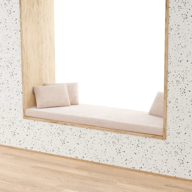 Ein gemütlicher Fensterplatz mit einem Rechteckigen Sitzfenster Polster nach Maß und zwei passenden Kissen ist in eine terrazzomäßig gemusterte Wand eingebaut und überblickt einen hellen Raum mit Holzfußboden.