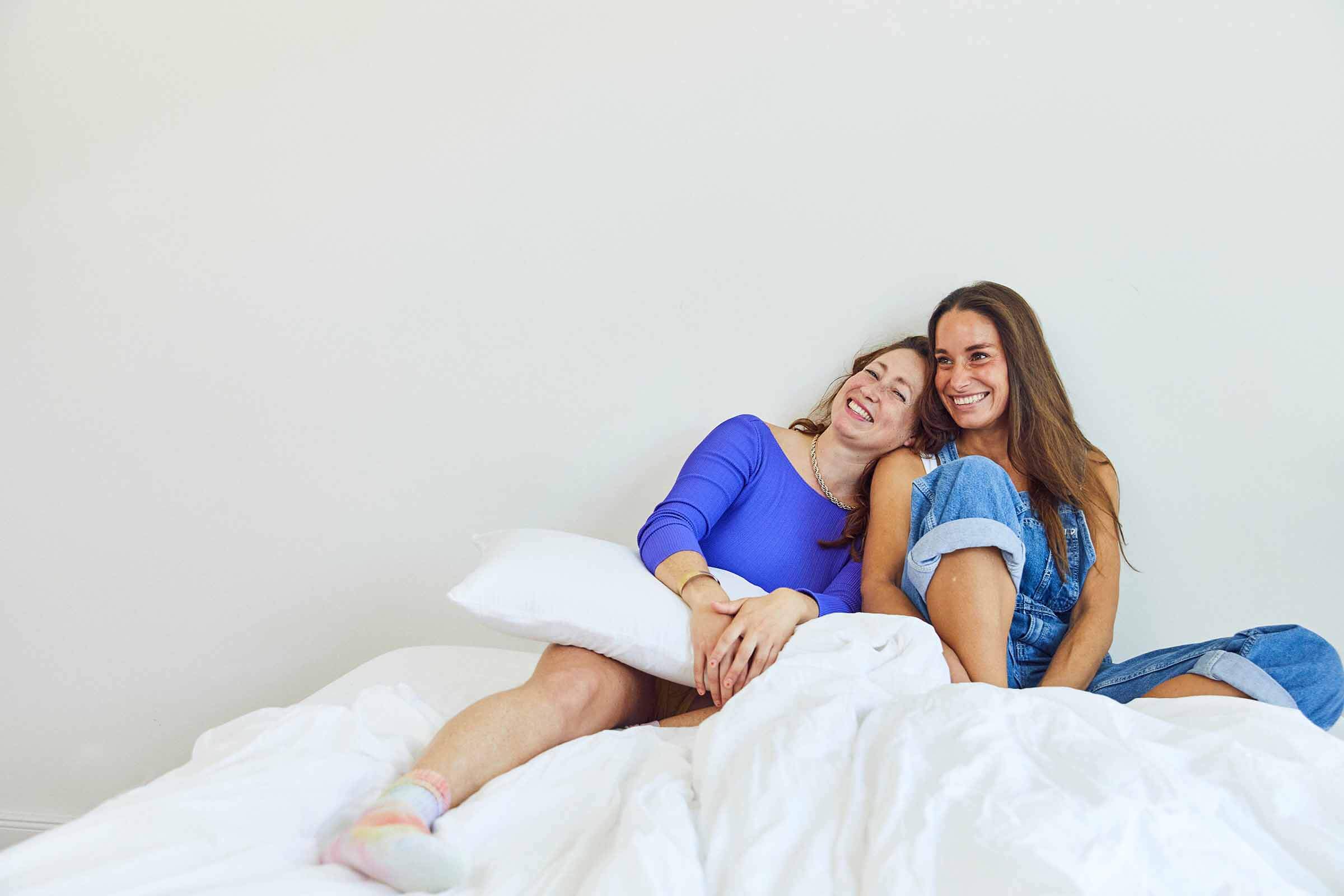 Zwei Frauen sitzen auf einem weißen Bett, lächeln und lehnen sich eng aneinander. Die eine trägt ein blaues Oberteil und hält ein Kissen, die andere trägt einen Jeans-Overall. Der Hintergrund ist eine schlichte weiße Wand.