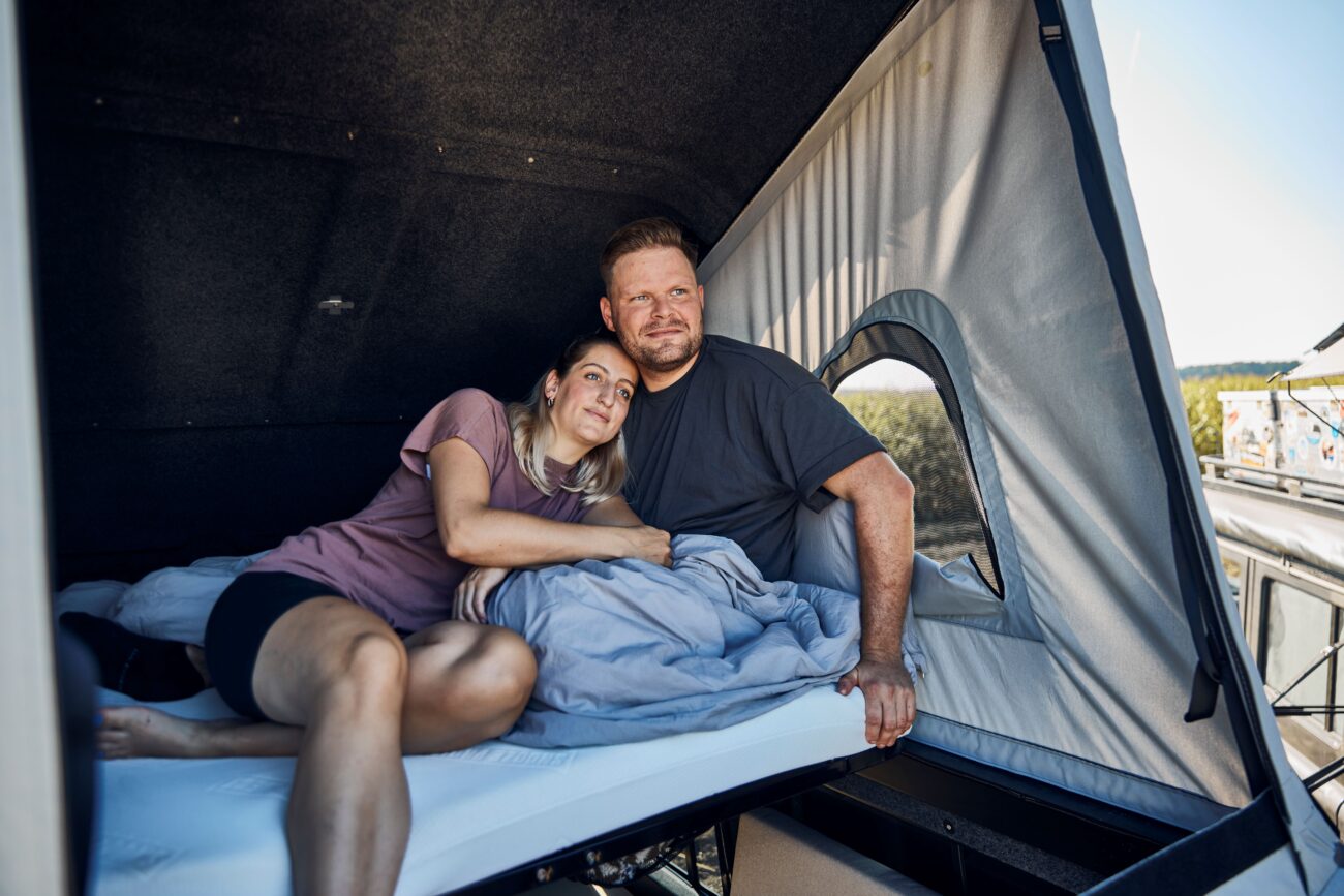 Ein Paar entspannt sich gemeinsam auf einem Bett in einem Wohnmobil, lehnt sich aneinander und lächelt. Das Sonnenlicht fällt durch das Fenster des Wohnmobils und beleuchtet die gemütlichen Betten und die bequeme Dachzeltmatratze, die um sie herum aufgebaut sind.