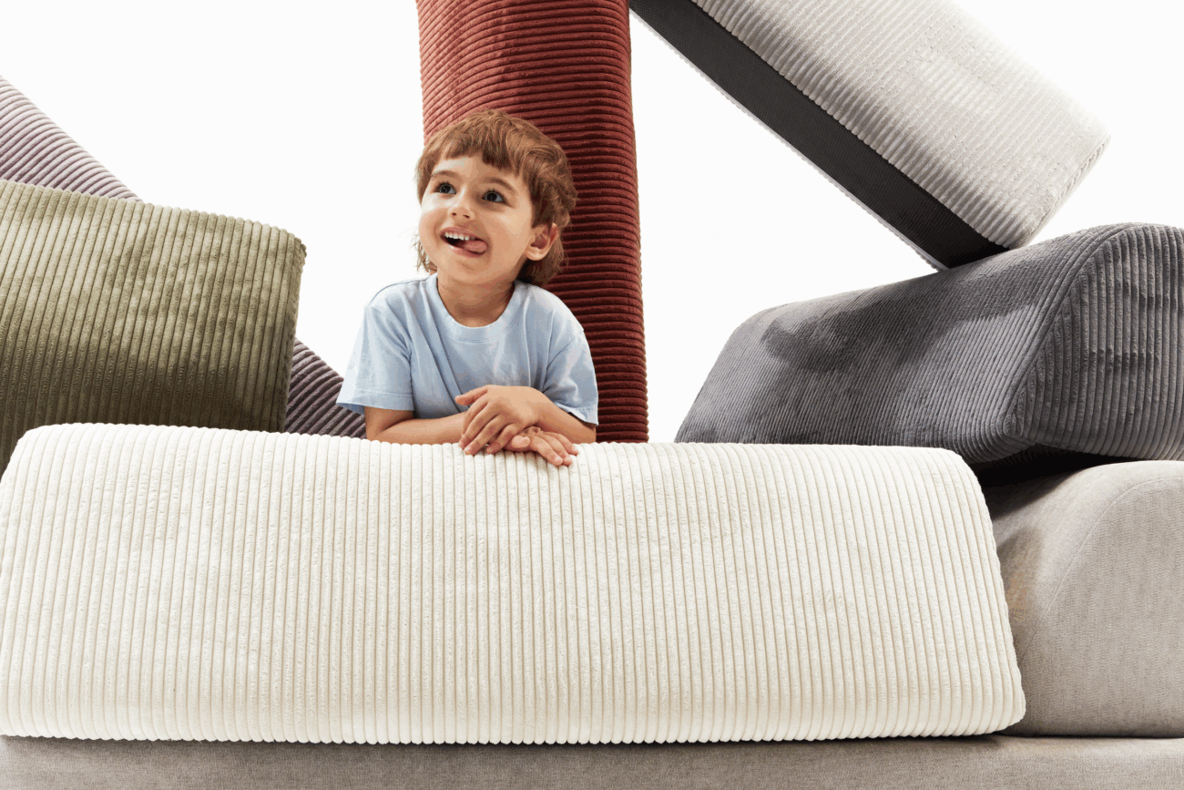 Ein lächelndes kleines Kind in einem hellblauen Hemd ist umgeben von großen, bunten, kordartigen Kissen, die in verschiedenen Formen und Richtungen angeordnet sind - eine ideale Inspiration für jede Schlafsofa-Kategorieseite. Der Hintergrund ist hell und weiß.