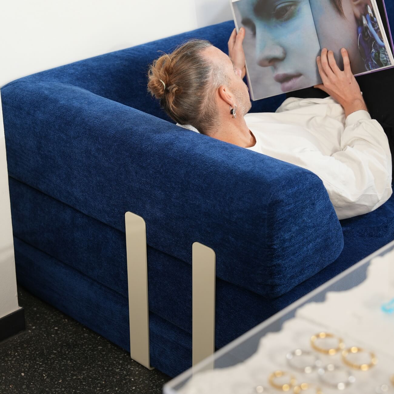 Eine Person mit Duttfrisur liegt auf einer dunkelblauen Couch, hält eine aufgeschlagene Zeitschrift mit einem Nahaufnahmeporträt in der Hand und schaut sie an. Auf einem Tisch im Vordergrund ist Schmuck zu sehen.