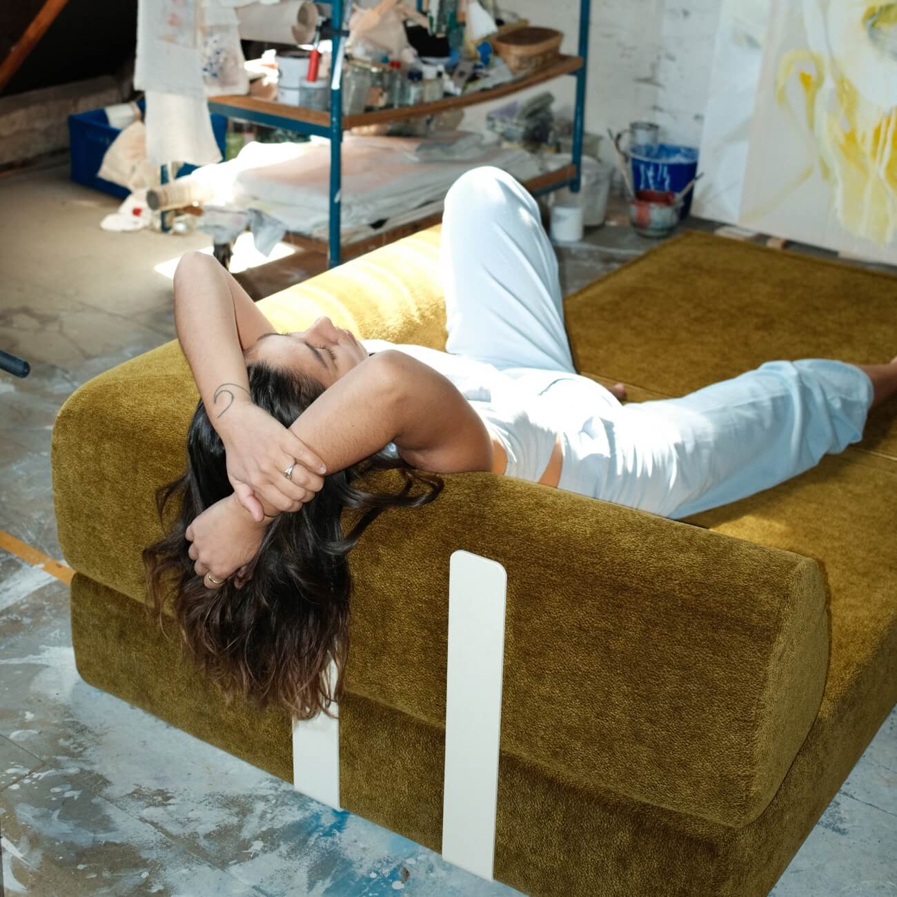 Eine Person mit langen Haaren und heller Kleidung entspannt sich auf einem senfgelben Sofa in einem sonnendurchfluteten, mit Kunstwerken gefüllten Atelier mit Regalen, Kunstzubehör und Leinwänden im Hintergrund.