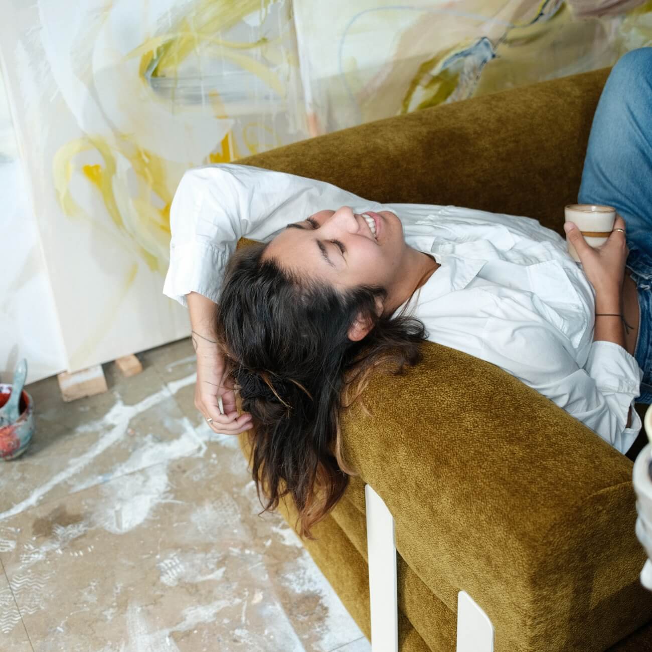 Eine Frau mit dunklem Haar lehnt lächelnd auf einem braunen Sofa und hält eine kleine Tasse. Im Hintergrund sind Kunstgegenstände und abstrakte Gemälde auf dem mit Farbe bespritzten Boden zu sehen.