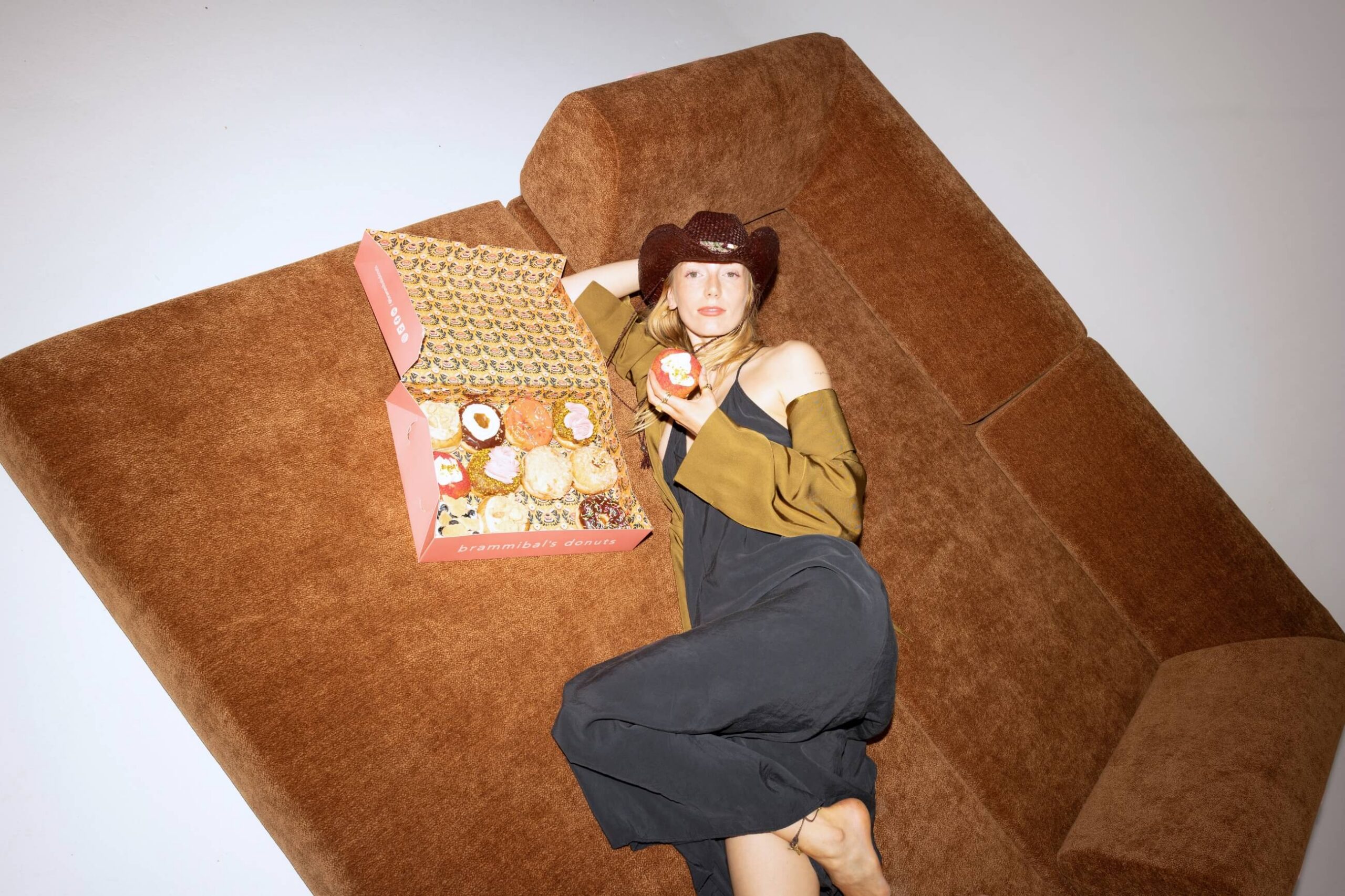 Eine Person in einem schwarzen Kleid und einem braunen Hut liegt barfuß auf einem braunen Sofa und hält einen Donut in der Hand. Neben ihr steht eine offene rosa Schachtel mit verschiedenen Donuts.