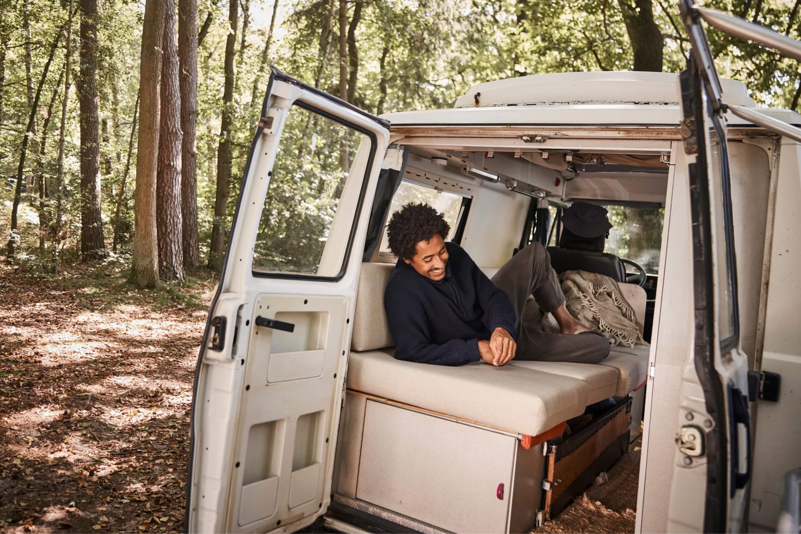 Eine Person entspannt sich auf einem Bett in einem weißen Wohnmobil, das in einem sonnenbeschienenen Wald geparkt ist, wobei die Türen des Fahrzeugs nach draußen geöffnet sind. Der Boden ist mit Laub bedeckt und das Sonnenlicht fällt durch die Bäume.