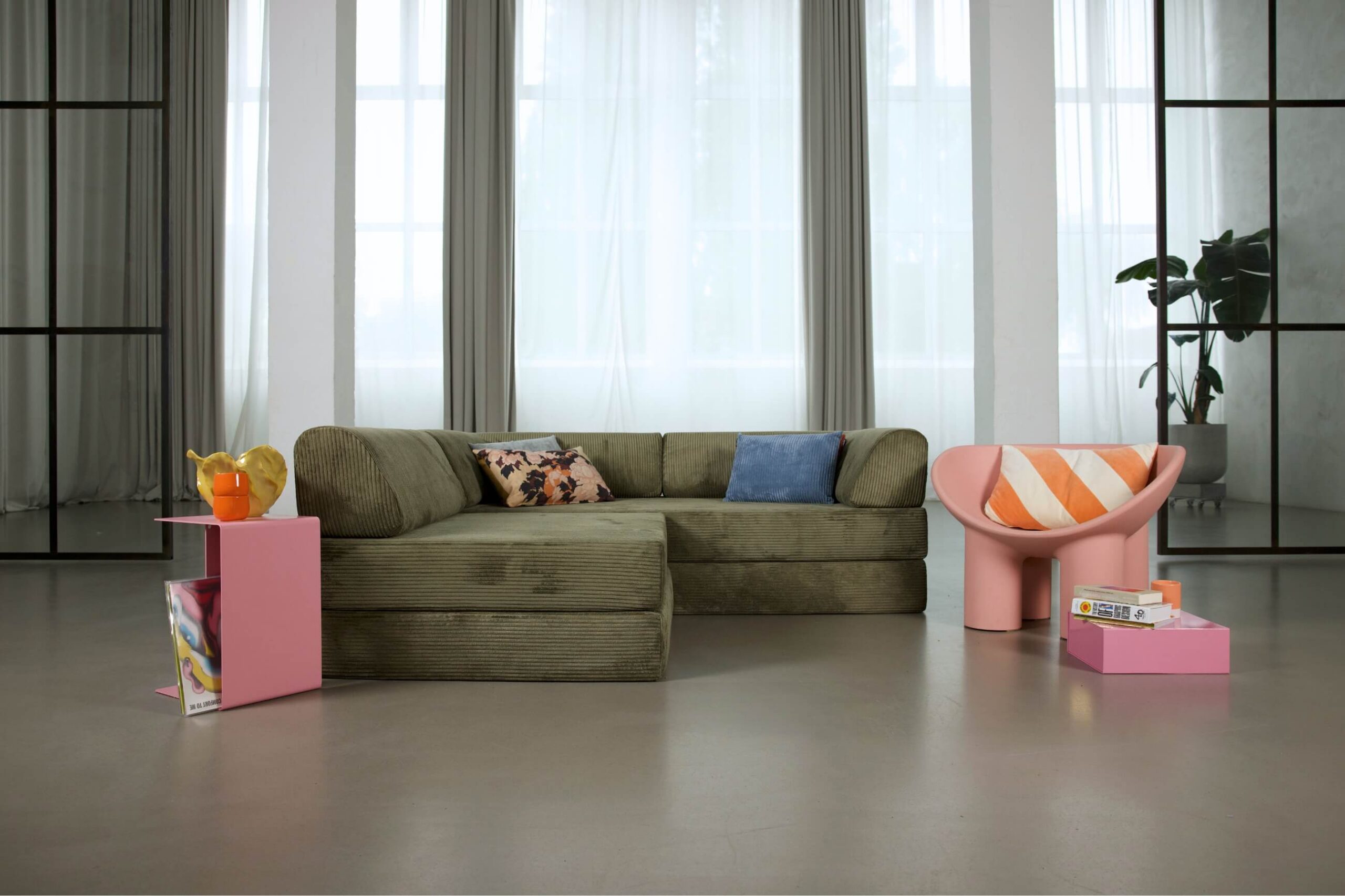Ein modernes Wohnzimmer mit einem grünen Sektionssofa, einem rosafarbenen Beistelltisch mit Obst und Zeitschriften, einem rosafarbenen Akzentsessel mit einem orange gestreiften Kissen, einem rosafarbenen Couchtisch mit Büchern und großen Fenstern mit transparenten Vorhängen.