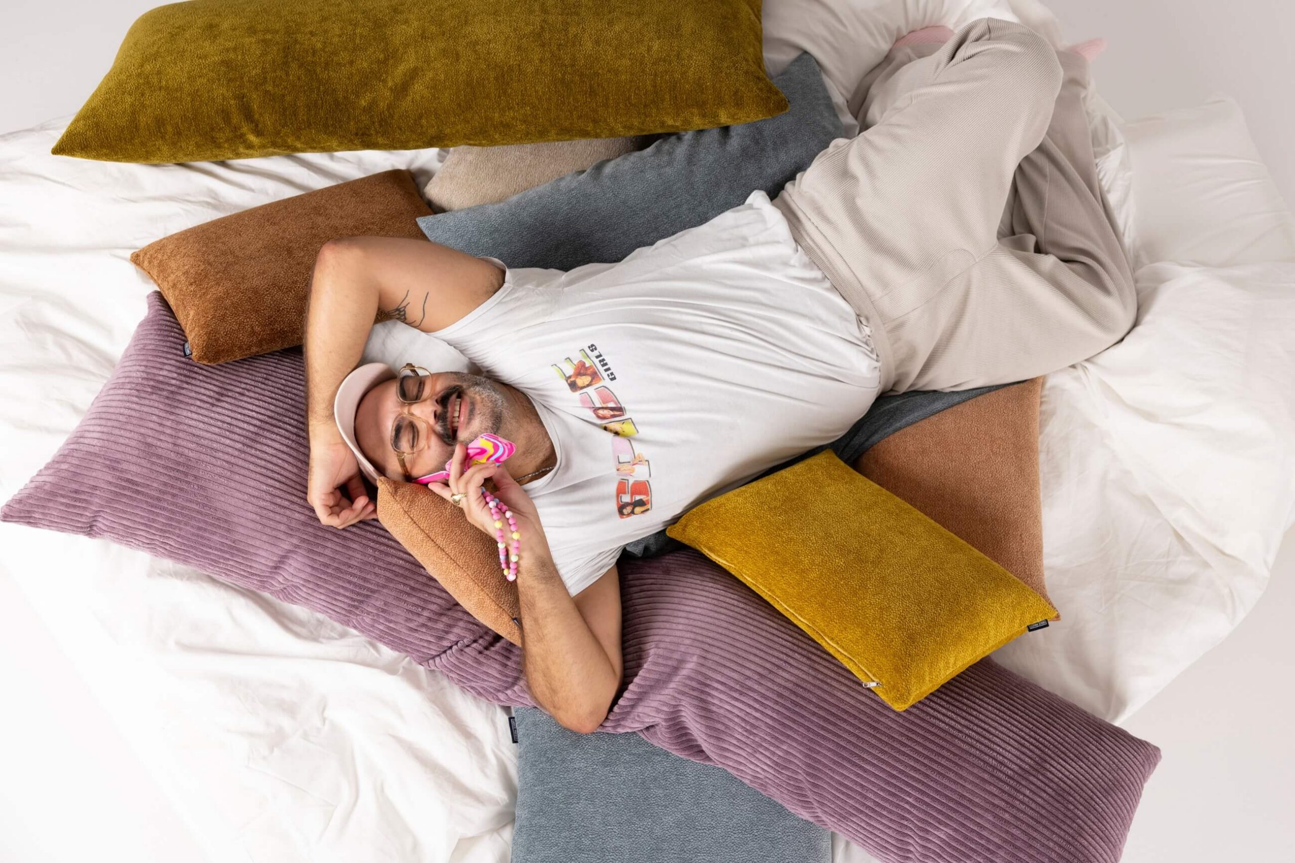 Eine Person in einem weißen T-Shirt und einer hellen Hose liegt auf einem Bett mit bunten Kissen, lächelt und telefoniert. Die Umgebung wirkt gemütlich und entspannt.