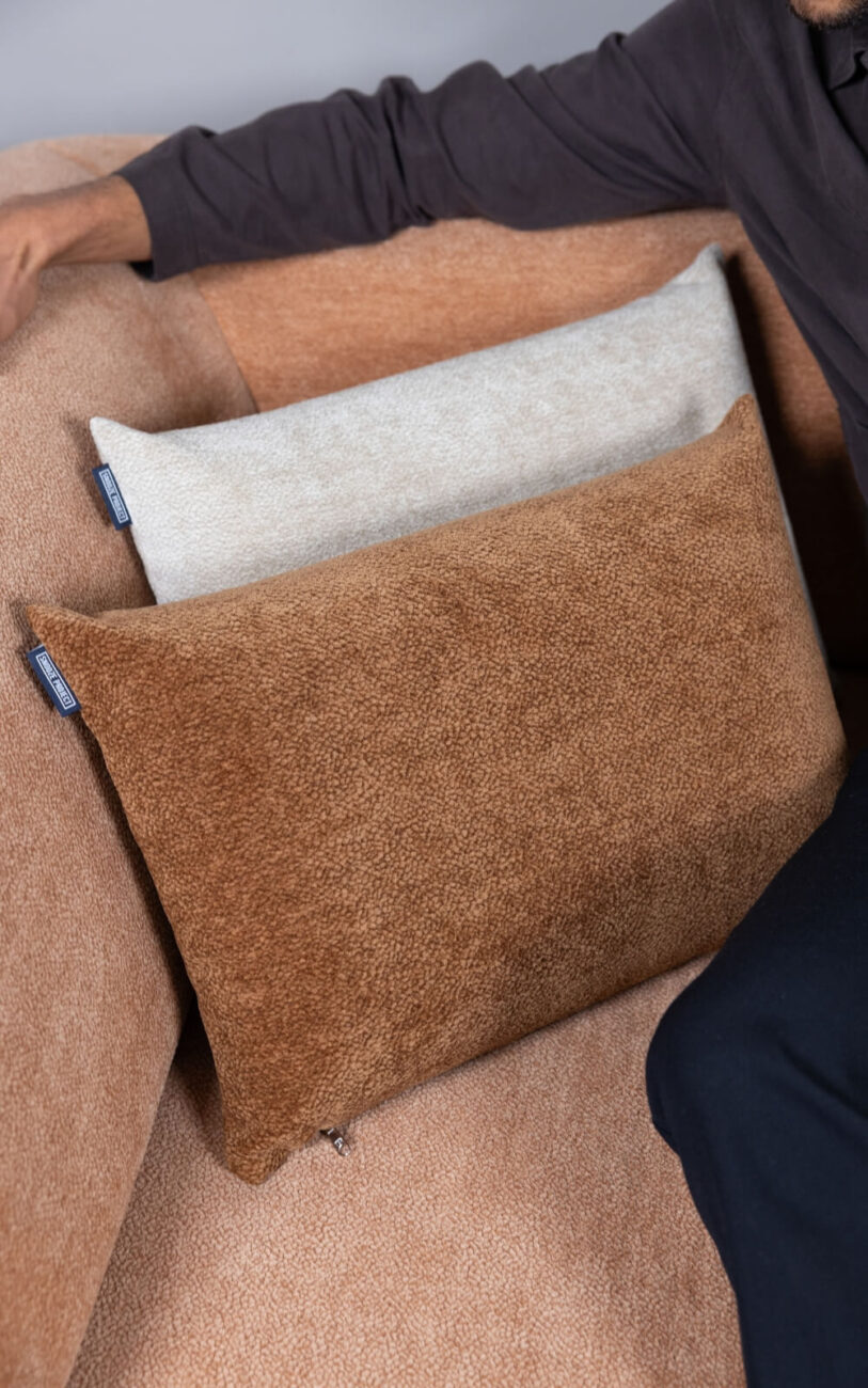 Eine Person sitzt auf einer beigen Couch mit zwei rechteckigen Kissen, einem hellbeigen und einem braunen, die auf dem Sitz ruhen. Man sieht den Arm der Person, die mit einem langärmeligen dunklen Hemd bekleidet ist.