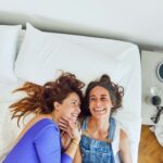 Zwei Frauen liegen auf einem Bett, lächeln sich an und schauen sich an. Die eine trägt ein blaues Oberteil, die andere einen Jeans-Overall. Sie wirken glücklich und entspannt, ihre Köpfe liegen eng beieinander auf dem weißen Bettzeug.