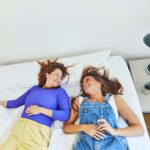 Zwei Frauen liegen auf einem Bett, stehen sich gegenüber und lächeln. Die eine trägt ein blaues Oberteil und eine gelbe Hose, die andere einen Jeans-Overall und ein weißes Hemd. Neben ihnen steht ein Nachttisch mit einer Brille und einer Lampe.