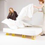 Zwei Personen machen ein Bett mit weißen Laken in einem hellen, minimalistischen Raum. Das Bett hat einen einfachen Holzrahmen mit gelben Beinen, und beide Personen heben die Laken zusammen, um sie über die Matratze zu legen.