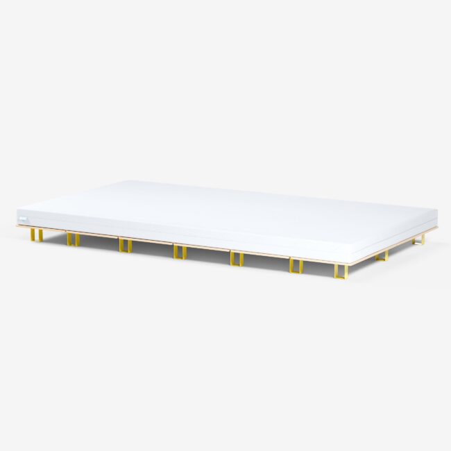Ein niedriges Bett mit einer weißen Matratze auf einem minimalistischen Holzrahmen, der von mehreren gelben Beinen getragen wird, vor einem schlichten hellgrauen Hintergrund.