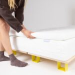 Eine Person, die einen dunklen Pullover und graue Socken trägt, hebt die Ecke einer weißen Matratzenauflage auf einem Bett mit einem Holzrahmen und hellgelben Beinen an.