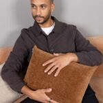 Ein Mann mit gestutztem Bart sitzt auf einer beigen Couch und trägt eine dunkelbraune Jacke über einem weißen Hemd. Er hält mit beiden Händen ein strukturiertes, hellbraunes Kissen über seine Brust und schaut zur Seite.