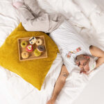 Ein Mann mit Brille und Schlafmaske liegt auf einem Bett mit einem gelben Kissen, neben dem ein Holztablett mit sechs verzierten Donuts steht. Er wirkt entspannt und ist mit einem weißen T-Shirt und einer beigen Hose bekleidet.