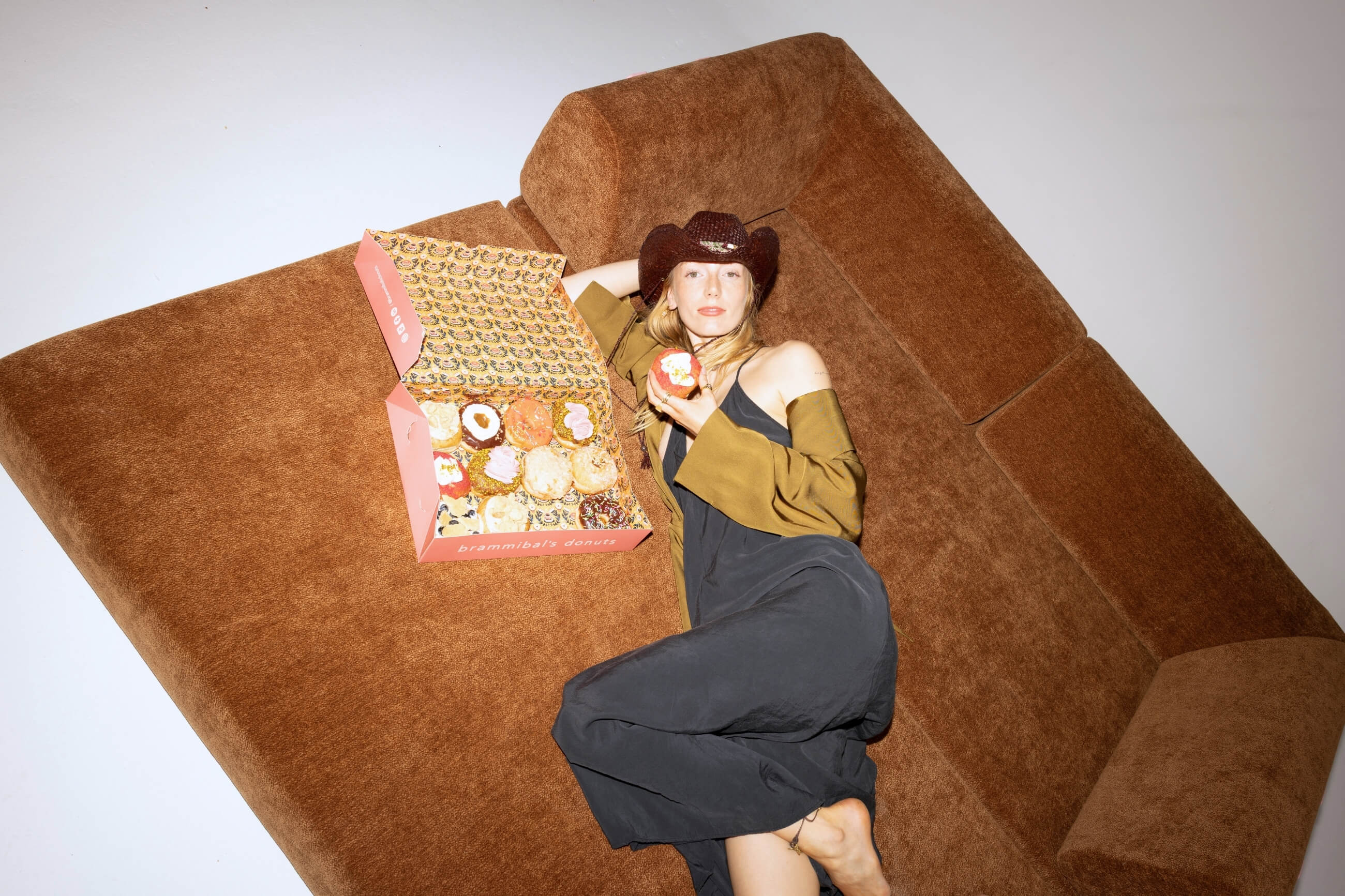 Eine Frau in einem schwarzen Kleid und einem braunen Cowboyhut liegt barfuß auf einem braunen Sofa und hält einen Donut in der Hand. Eine rosa Schachtel mit verschiedenen Donuts liegt neben ihr auf dem Sofa.