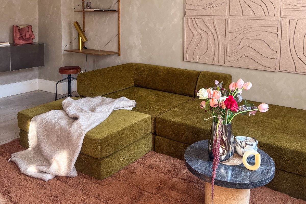 Ein modernes Wohnzimmer mit dem Levi-Sofa in Grün, das mit einer beigen Decke überzogen ist. Strukturierte Wandkunst, UGC-inspiriertes Dekor und ein schicker Beistelltisch mit Marmorplatte verstärken die gemütliche Atmosphäre auf einem weichen, rosafarbenen Teppich.