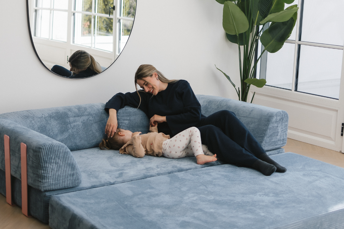 Eine Frau und ein Kind entspannen sich und lächeln gemeinsam auf einem blauen Levi-Sofa in einem hellen, modernen Wohnzimmer mit großen Fenstern, einem runden Spiegel und einer hohen Grünpflanze - das perfekte UGC für gemütliche Wohninspiration.