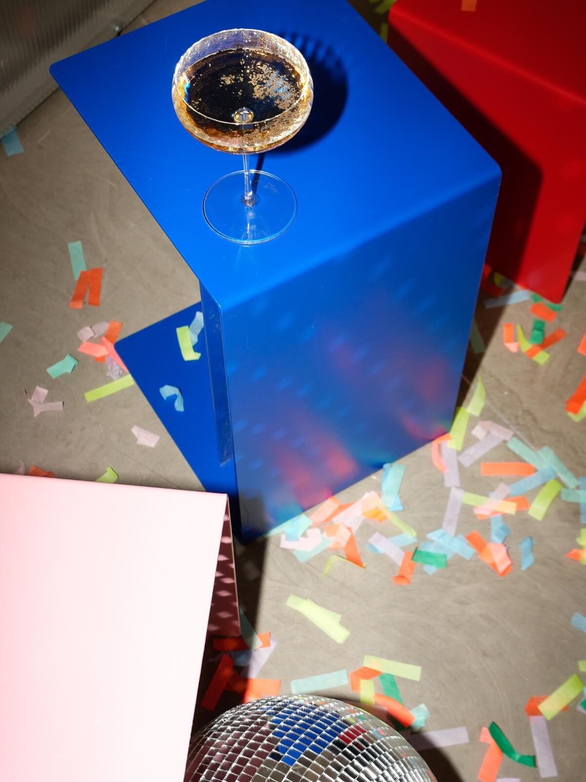 Auf einem hellblauen Tisch steht ein Glas mit einem dunklen Getränk. Auf dem Boden liegen bunte Konfetti verstreut; dort sind außerdem ein rosa Tisch, ein roter Tisch und ein Teil einer Discokugel zu sehen.